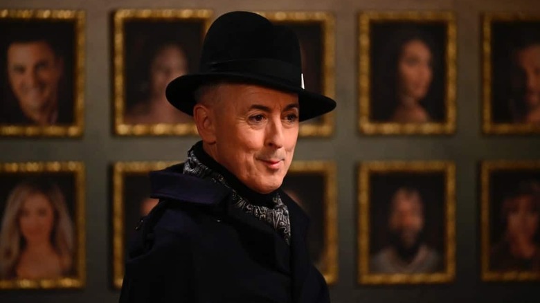 Alan Cumming in a stylish black ensemble in front of a wall of contestant portraits