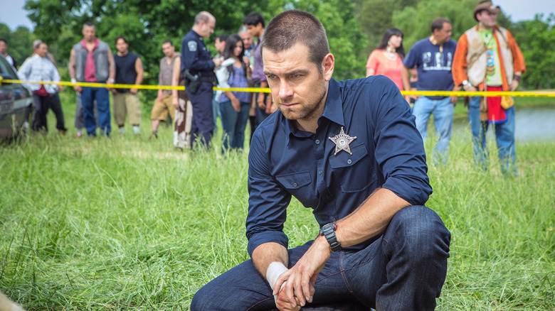 Lucas, wearing sheriff's badge crouching in the grass at crime scene in Banshee