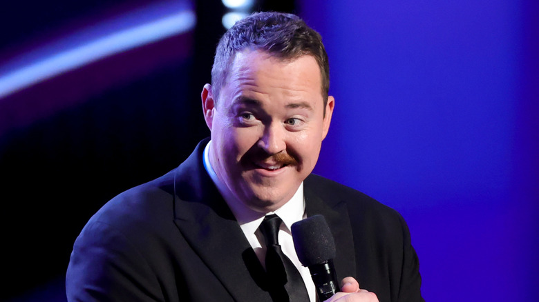 Shane Gillis in a suit holding a microphone