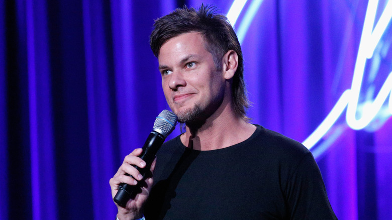 Theo Von holding a microphone in front of blue curtains