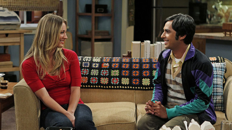 Penny and Raj sitting on her couch smiling and talking on The Big Bang Theory