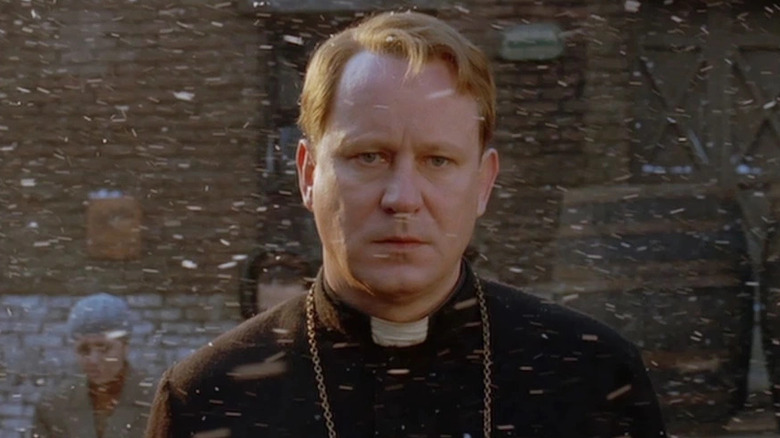 Merrin stands in the snow in front of a brick building in Dominion: A Prequel to the Exorcist