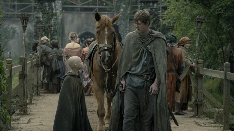 Ser Duncan, wearing a green tunic, looks down at the small Egg, bald with a grey tunic, while walking their horse through the roads of Westeros in "A Knight Of The Seven Kingdoms"