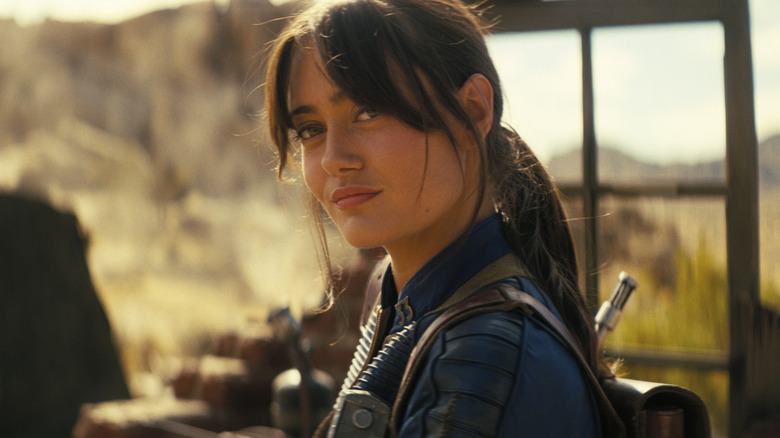 Lucy stands by a door smirking in the desert in 