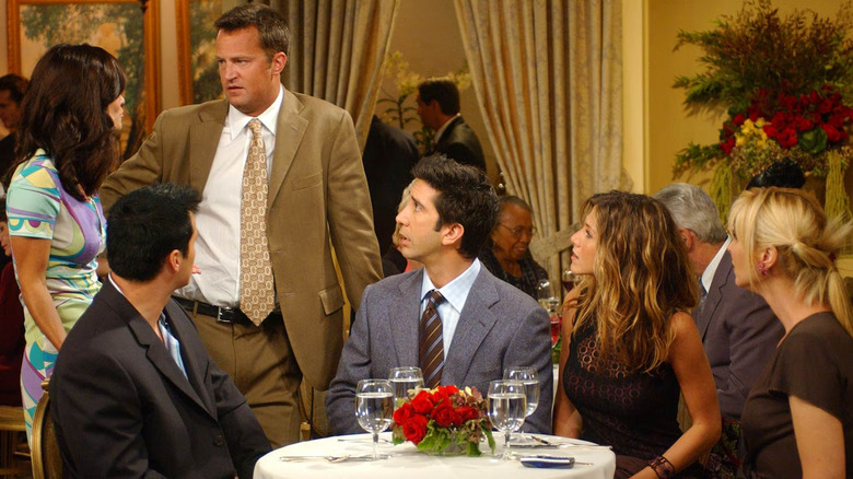 Monica and Chandler arriving late to dinner with Joey, Ross, Rachel, and Phoebe sitting at a small round table in Friends