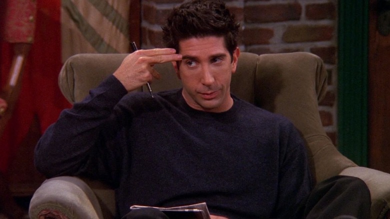Ross sitting in a Central Perk armchair holding two extended fingers up to his temple in Friends