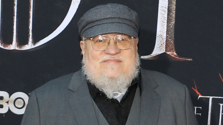 George R. R. Martin smiles in front of a promotional poster for Game of Thrones