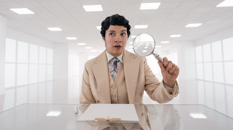 Muriel sits at a desk in heaven holding a magnifying glass in "Good Omens 3"