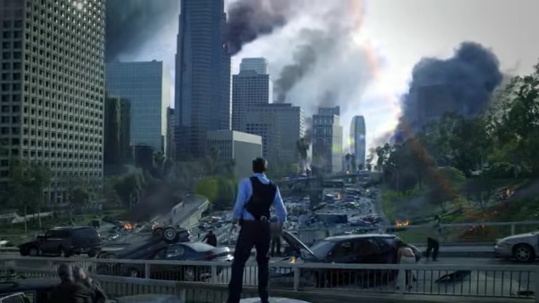 A man in a bulletproof vest looks out over a ruined city full of crashed cars and smoking buildings, in FlashForward