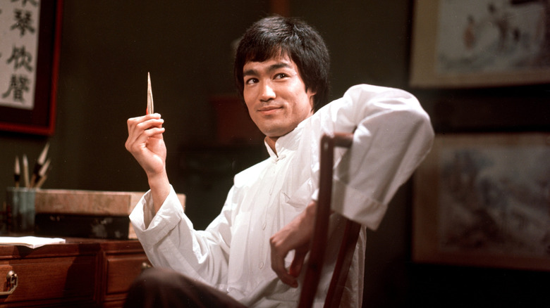 Lee holding up a pen at his desk and smiling (1973)