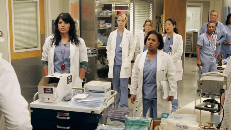 Sara Ramirez as Callie Torres, Chandra Wilson as Miranda Bailey, and Sandra Oh as Cristina Yang among others stand in a hospital in Grey's Anatomy.