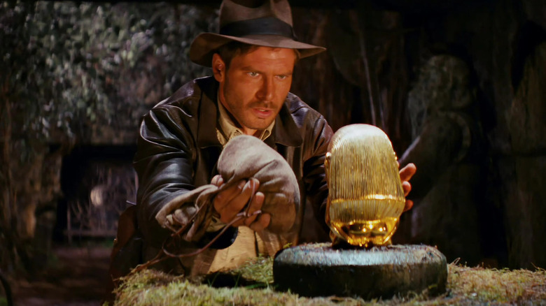 Indiana Jones holds a bag while reaching for a gold statue in Raiders of the Lost Ark (1981)