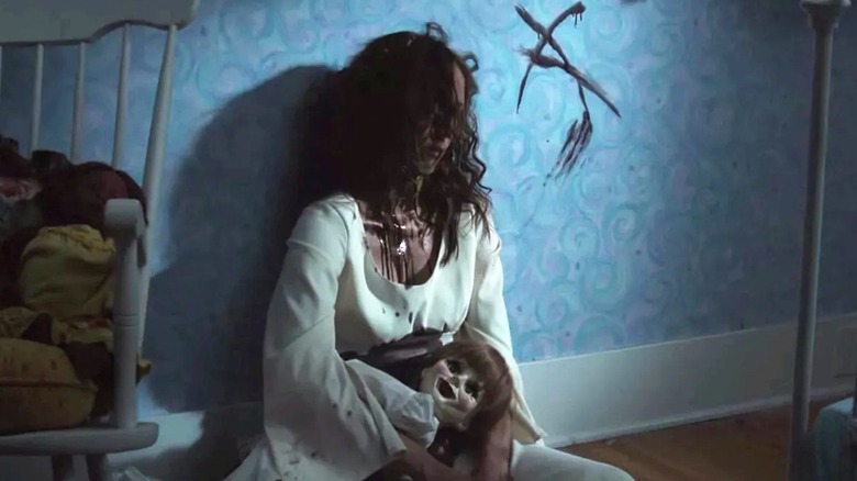 A dead Annabelle leaning against a wall, holding the porcelain doll, in Annabelle