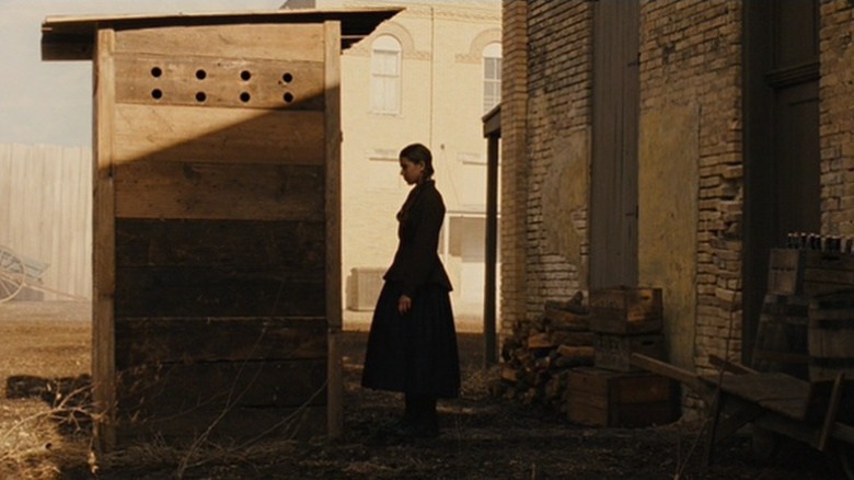 Mattie Ross standing in front of an outhouse
