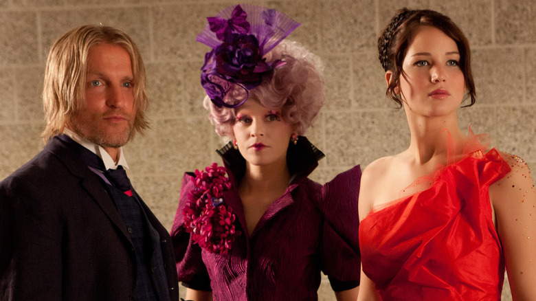 Effie, Haymitch, and Katniss stand in front of a wall