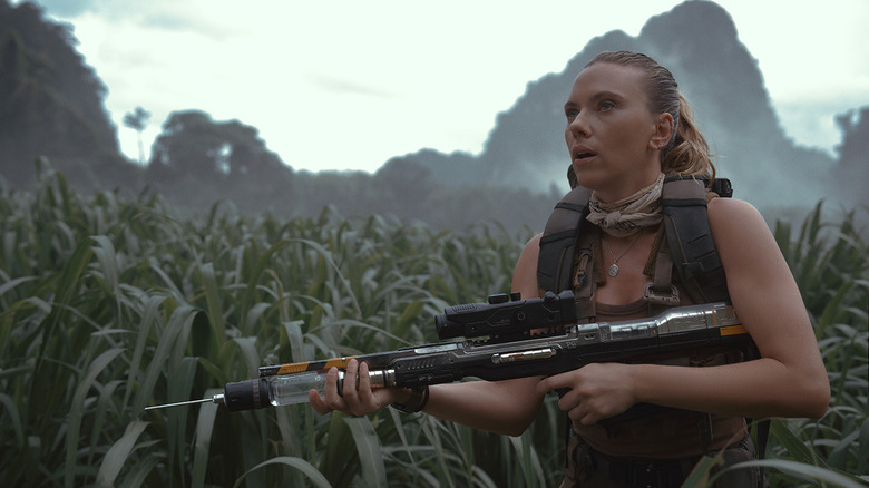 Zora Bennett walking with big needle gun through tall grass