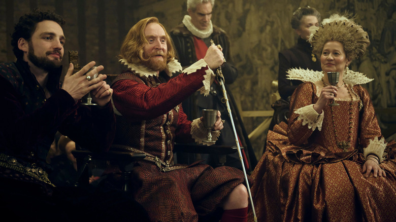 Royal court members cheer and raise glasses as the King holds a sword in "Mary & George" (2024)