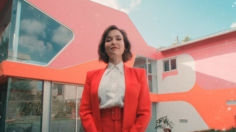 A woman in an orange suit standing outside a colorful house in "Give Me an A."