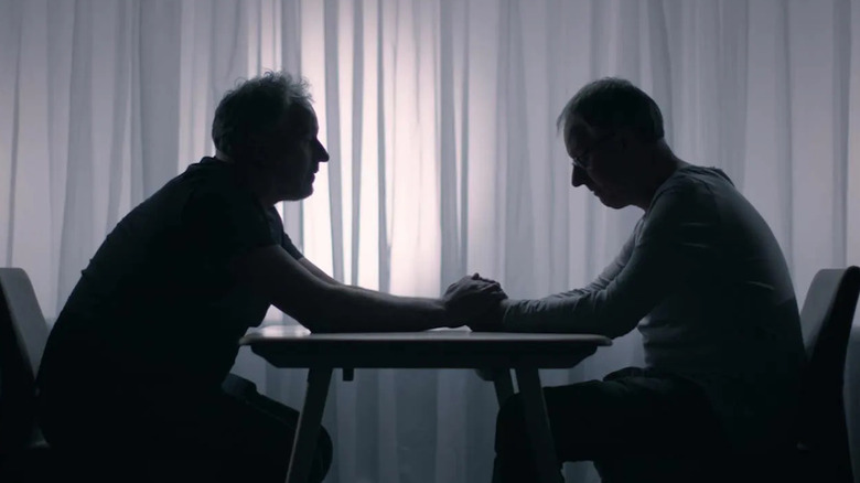 Marcus and Alex sitting in darkness at a table holding hands in Tell Me Who I Am