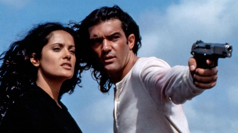 El Mariachi holding up a pistol while wearing a white shirt as Carolina watches beside him in Desperado