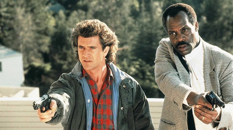 Martin Riggs holds a pistole while wearing a jacket and red flannel shirt as Roger Murtagh stands beside him in a suit with a gun in "Lethal Weapon"