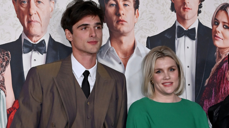 Jacob Elordi and Emerald Fennell stand in front of a "Saltburn" poster