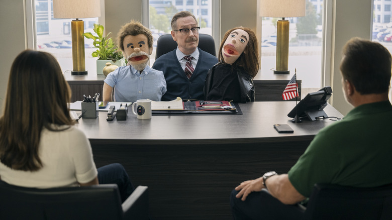 A CIA operative sitting and holding two puppets as he faces and talks to a man and a woman