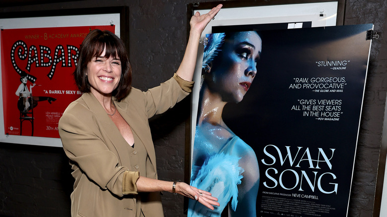 Neve Campbell smiling showing off Swan Song poster