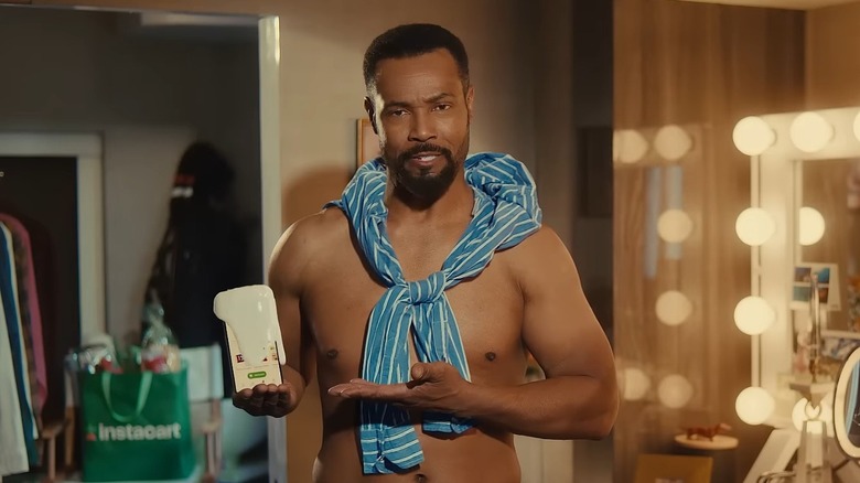 Isaiah Mustafa in a trailer in character as the Old Spice guy, his phone gushing foam, an Instacart tote bag visible behind him
