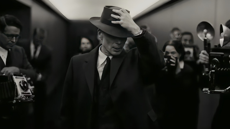 Oppenheimer holding his hat as he steps through crowd of photographers
