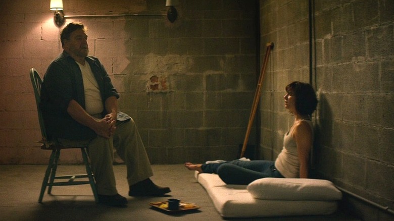 Michelle sits on a mattress in a basement while Howard sits in a chair talking to her in 10 Cloverfield Lane (2016)