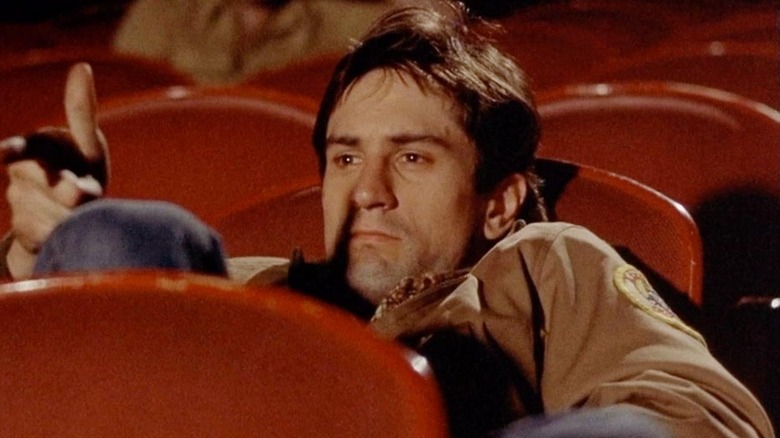 Travis Bickle sits in a movie theater and points a finger gun at the screen in 