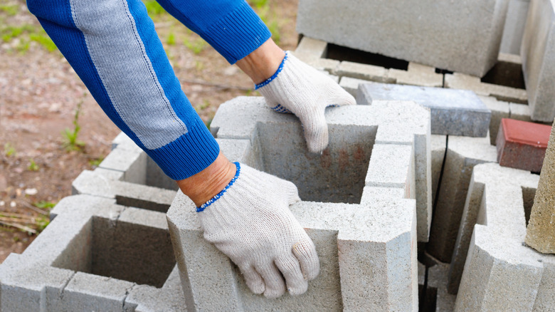 A person wearing cloth gloves picks up a large hollow concrete block with two hands.
