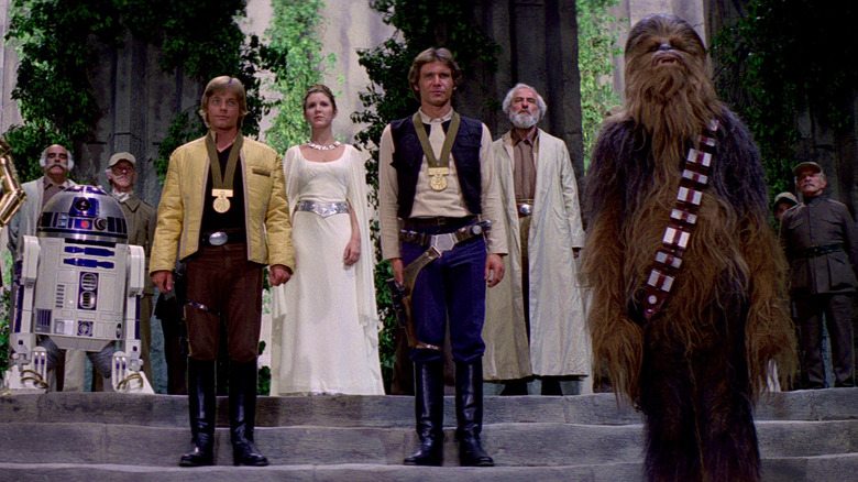 Luke Skywalker, in a yellow jacket, and Han Solo, in a blue vest, wear gold medals as they stand in front of Princess Leia and the other rebel forces in "Star Wars Episode 4: A New Hope"