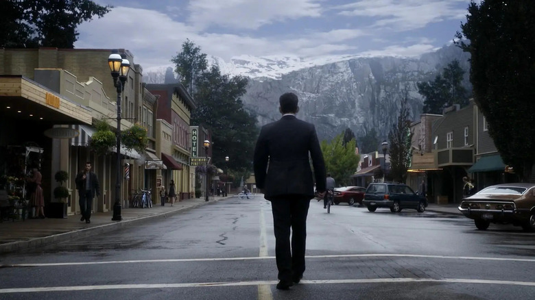 Ethan Burke walks down the middle of the street with his back turned while wearing a suit amidst the mountain town in 