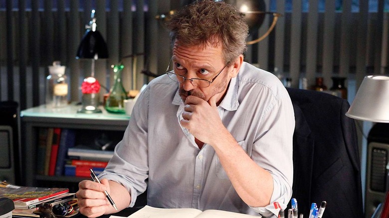 House at his desk wearing glasses looking stressed on House