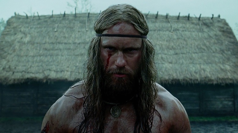 Amleth, with blood on his face, stands in front of a hut in "The Northman" (2022)