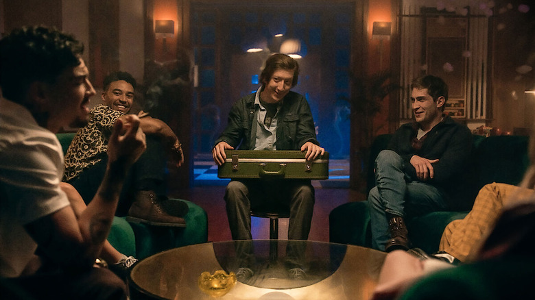 The friends sit around a table and laugh, while Forbes holds a suitcase in his lap in It's What's Inside
