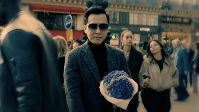 Blind assassin Caine standing in a crowd in Paris holding a bouquet of blue flowers