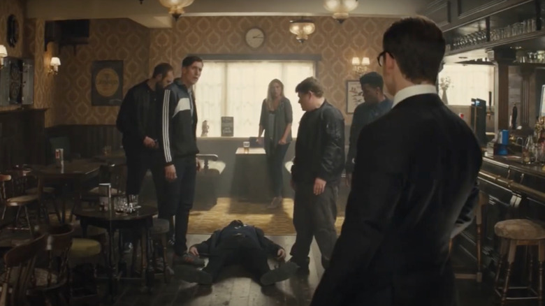 Eggsy (Taron Egerton) standing, facing his mother and his stepfather's crew after knocking out his stepfather (Geoff Bell) with a glass tumbler