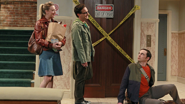 Penny carrying groceries standing next to Leonard talking to Sheldon sitting on the floor on The Big Bang Theory