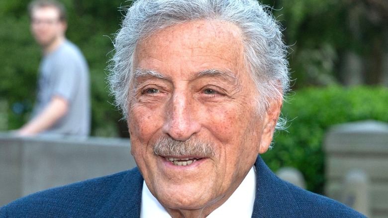 Tony Bennett standing outside in a suit with a mustache with someone staring at him
