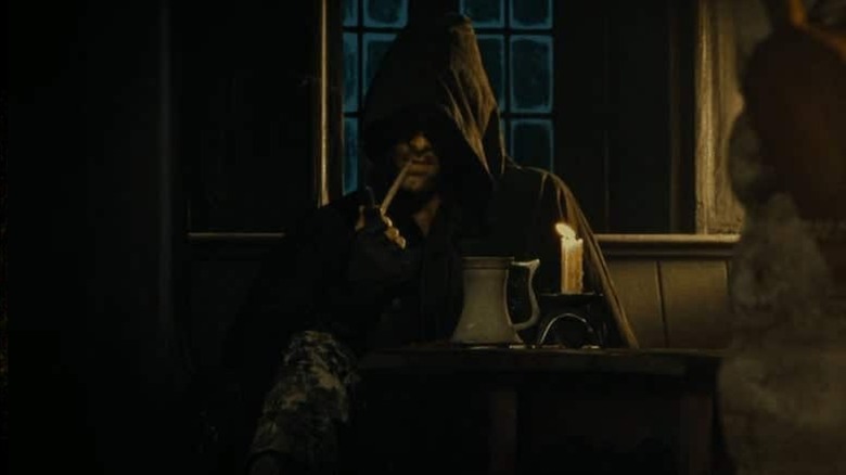 Aragorn sits at a table in a Ranger's cloak, smoking a pipe