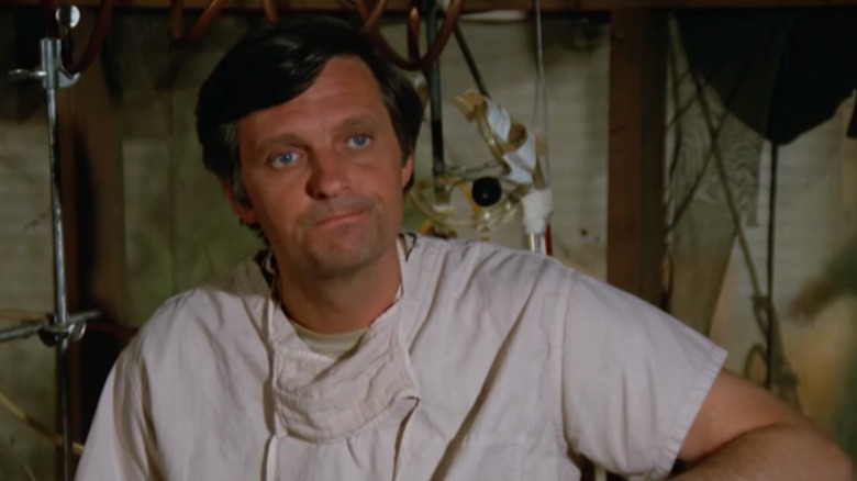 Alan Alda as Hawkeye Pierce, seated and smirking