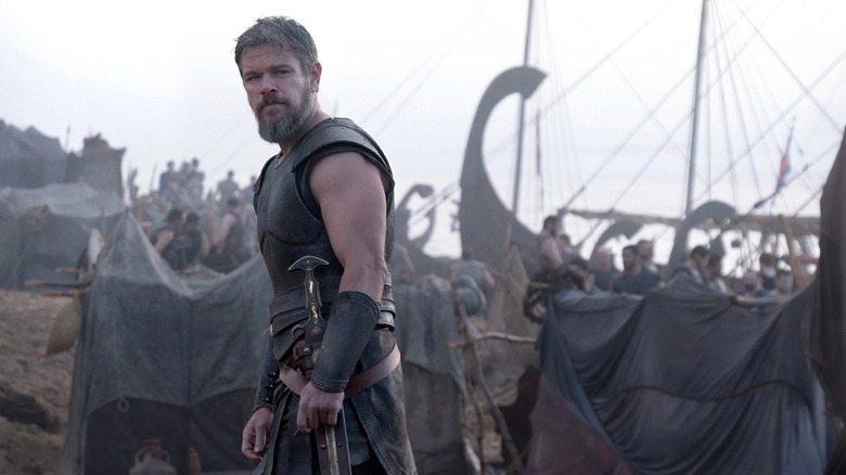 Odysseus looking over his shoulder on a beach while men prepare ships for launch in the background in The Odyssey (2026)