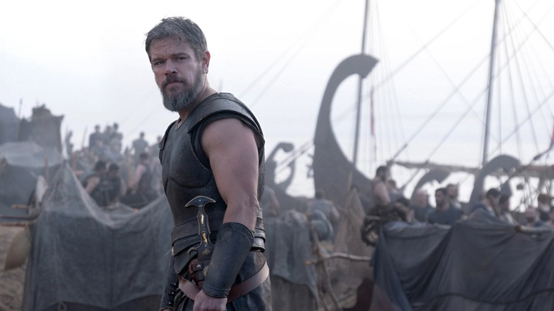 Odysseus looking over his shoulder on a beach while men prepare ships for launch in the background in The Odyssey (2026)