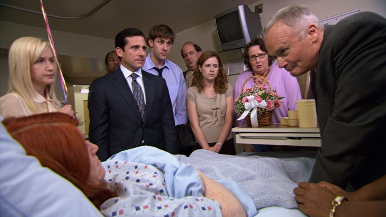 The employees of Dunder Mifflin gather around a hospital bed