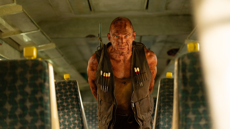 Dr. Kelson, coverded in orange iodine and a green vest, leans over with his hands behind his back inside a train compartment in 