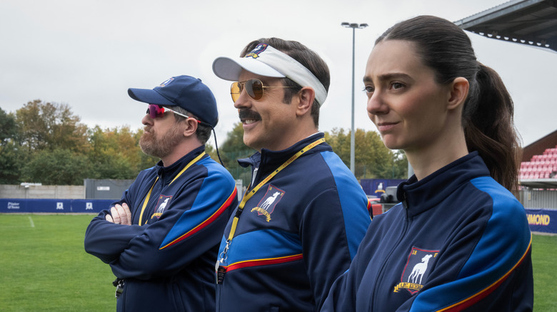 Beard, Ted, and a female coach played by Tanya Reynolds stand next to each other on the field at Nelson Road in Ted Lasso season 4 (2026)