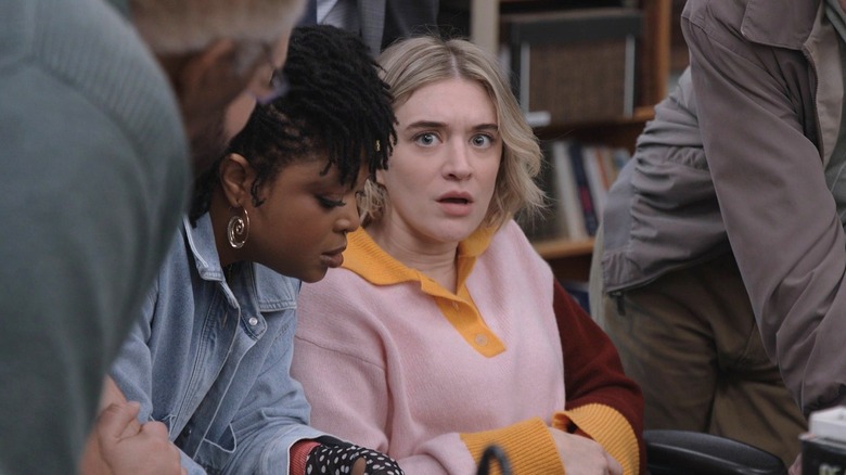 Adelola (Gbemisola Ikumelo) looking at Mare's desk while Mare (Chelsea Frei) is looking at the camera surrounded by other coworkers